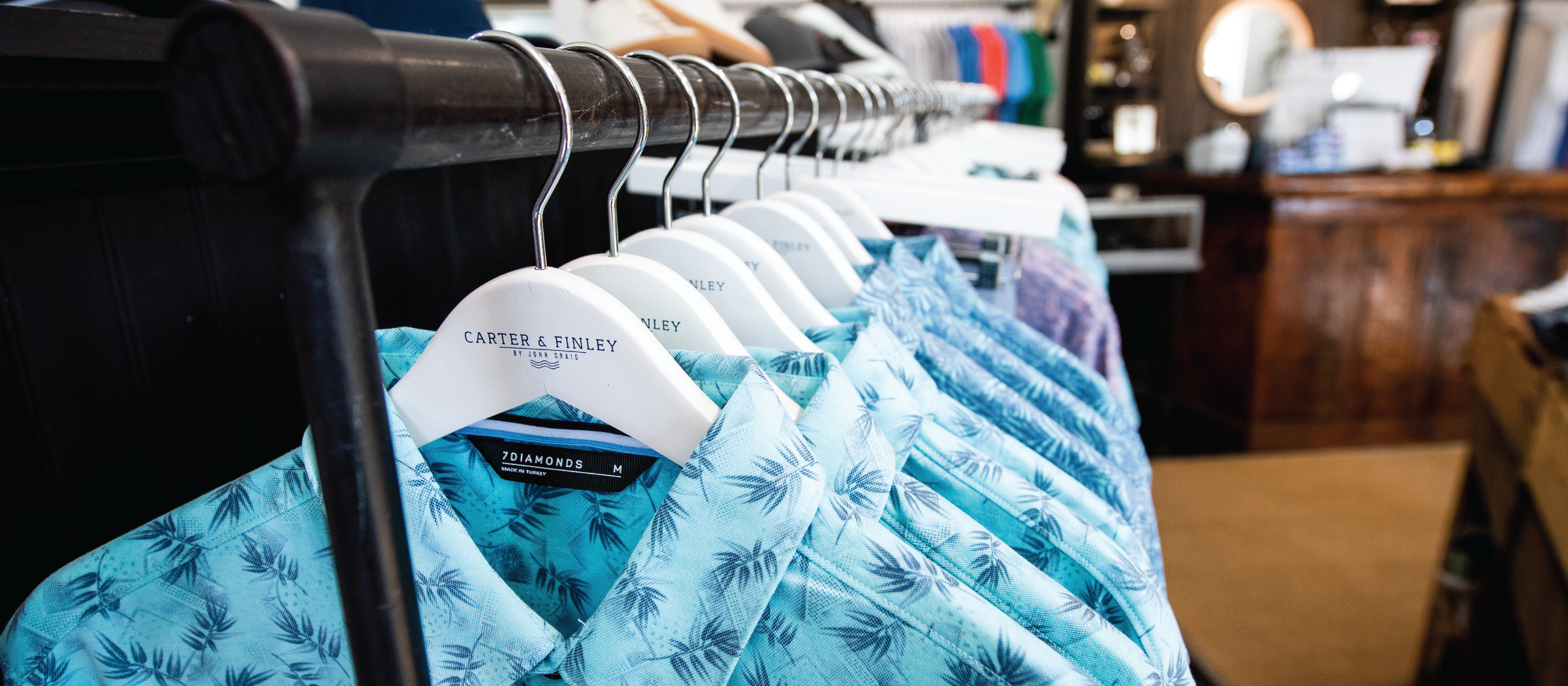 A row of blue, tropical-patterned shirts hanging on a clothing rack. The shirts, neatly arranged on white hangers, convey a beachy, relaxed vibe.
